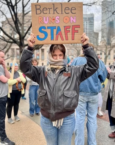 A gathering of Berklee staff members in front of Christian Science Plaza in Boston. In the foreground, a staff member holds up a handmade cardboard sign reading "Berklee runs on staff".